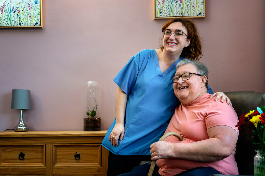 Carer standing hugging older man with walking stick sitting down. Both are smiling at the camera warmly