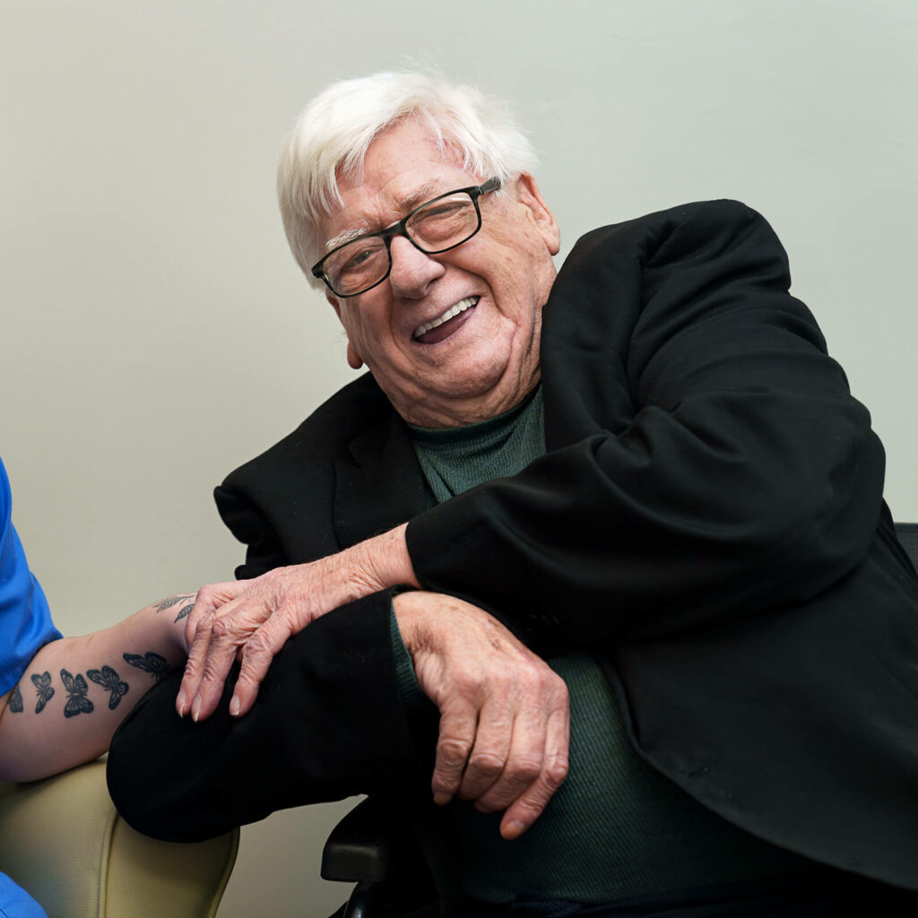 Older man laughing and looking at the camera, holding the hand of a carer just out of shot