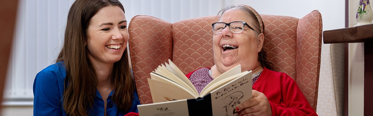 An older woman sits in an armchair smiling as a young woman reads her a book